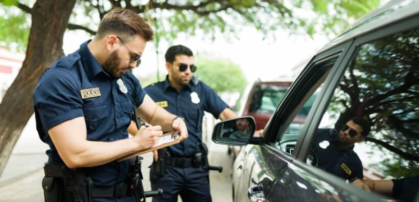 Police officers issuing a fix-it ticket to a driver during a roadside traffic stop, with one officer writing on a notepad beside the vehicle.