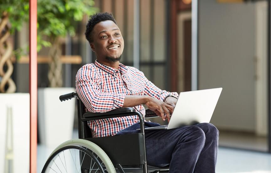 smiling african american man sitting in wheelchair and using laptop