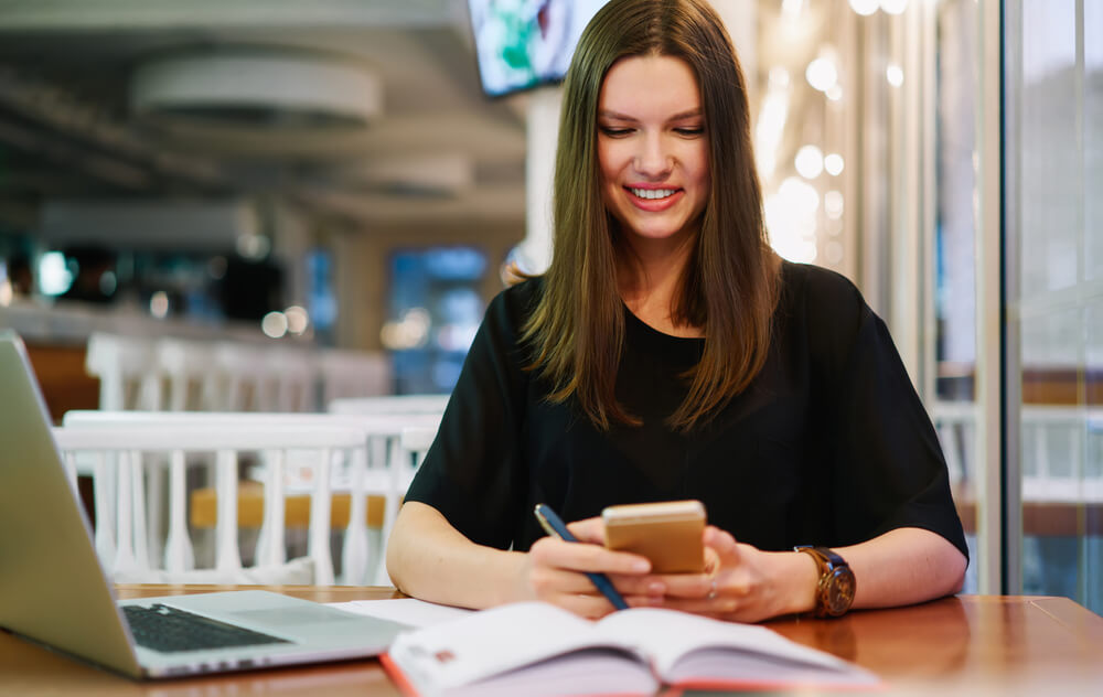 adult woman sitting at modern cafe with laptop and messaging via smartphone while holding pen