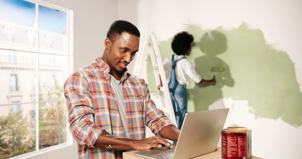 happy man with laptop with his wife painting the wall in the background