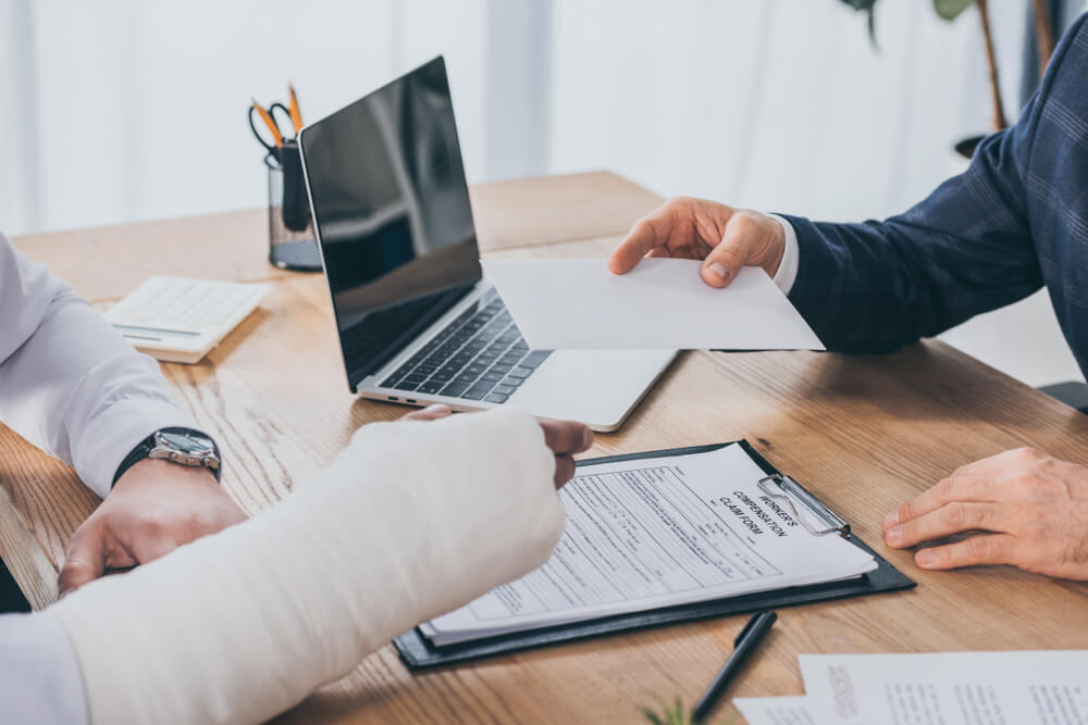 cropped view of businessman sitting at table and giving sheet of paper to workers with broken arm in office, compensation concept