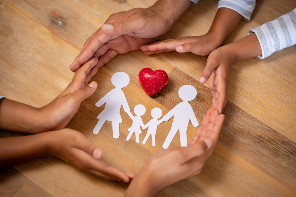 Close up hands of father, mother and daughters protecting family paper cutout with red heart. Hands of family with paper cutting and red heart shaped symbol stone. Unity, insurance and love concept.