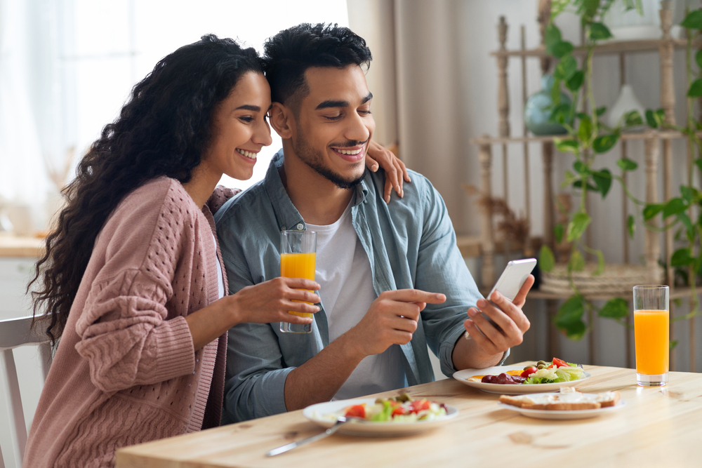 Cheerful,Arab,Spouses,Using,Smartphone,While,Eating,Breakfast,In,Kitchen, young couple eating breakfast together at table