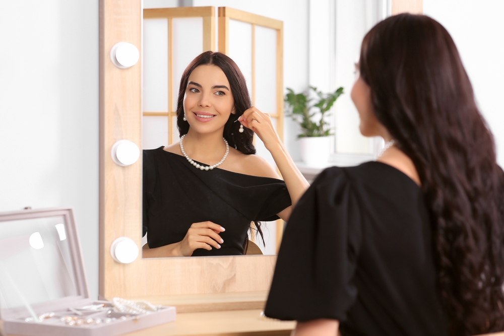 Young woman trying on elegant pearl earring near mirror indoors