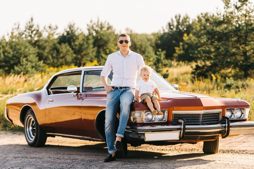 Father and son sit on the hood of a classic car in the sun