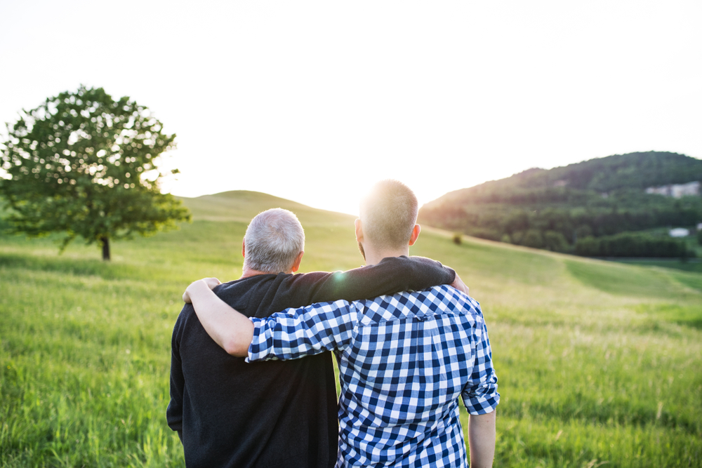 A father stands with his son with their arms around each other backs to camera