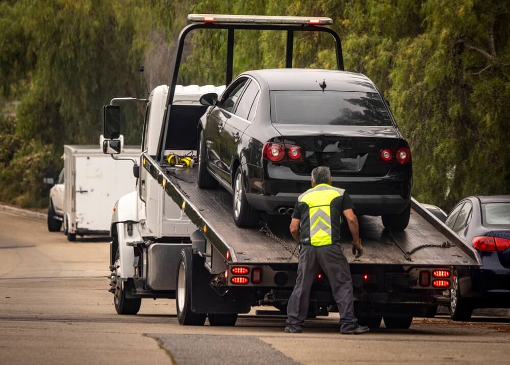 Roadside assistance worker loading a disabled car onto a tow truck on a tree-lined road, illustrating a situation in which people consider whether to tip roadside assistance.