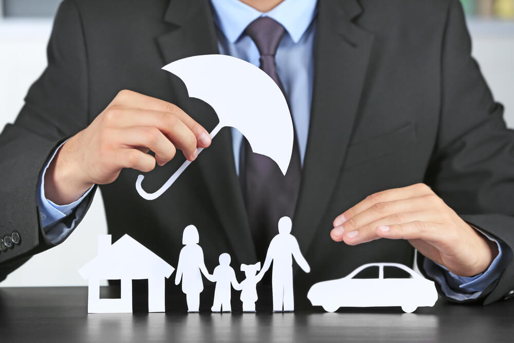 A male insurance agent from the neck down holding a cutout of an umbrella over cutouts of a home, family and car.