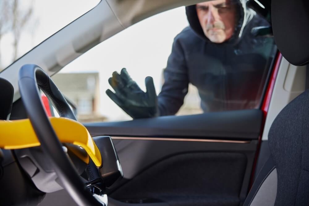 A man in a hooded jacket peers into a locked vehicle protected by a steering wheel lock, showing how anti-theft devices for cars help deter theft attempts.