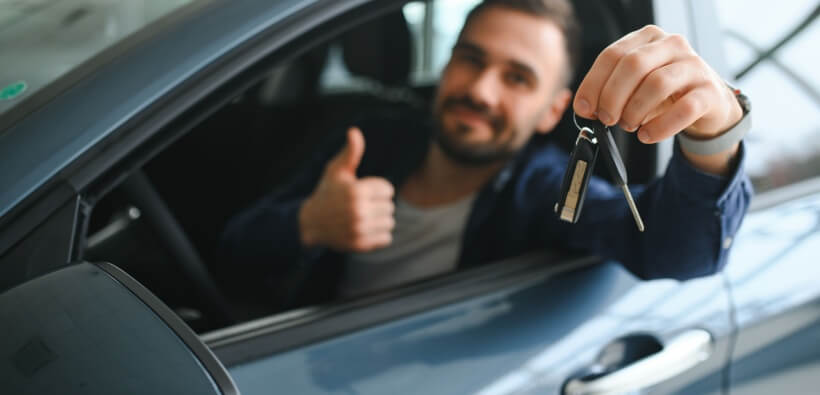 Driver in a car holding up rental car keys and giving a thumbs up, representing a positive rental car experience and vehicle pickup.