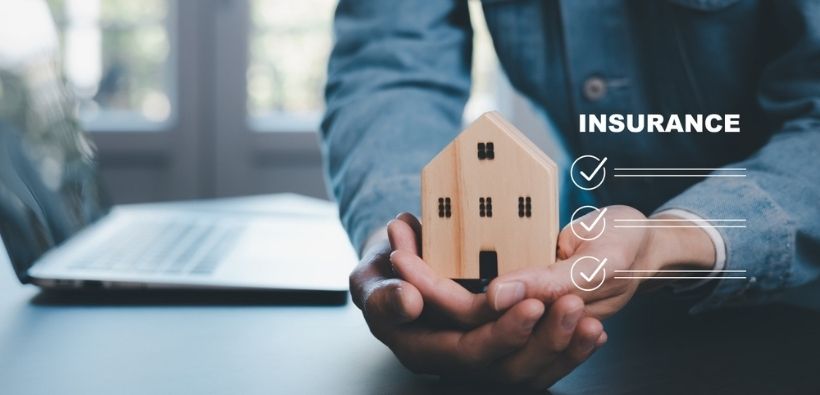 Hands holding a small wooden house with insurance checklist graphics, helping explain understanding replacement cost vs. actual cash value in home insurance.