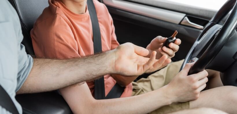 Parent handing car keys to a teenager inside a vehicle, illustrating the process of adding a teen driver car insurance policy.