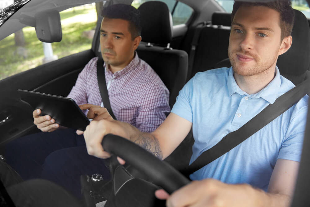 Young male driver with driving course instructor in the passenger seat, taking notes. How taking a driving course can lower car insurance rates.