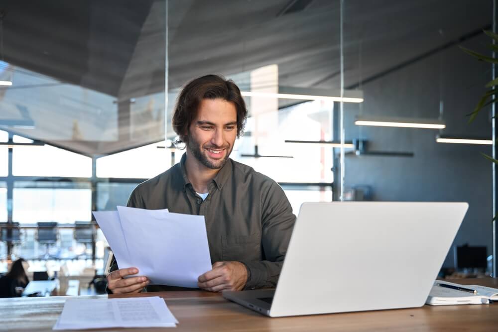Happy busy young business man accountant reading file report documents checking data online looking at laptop computer paying financial loan, analyzing the best business insurance.