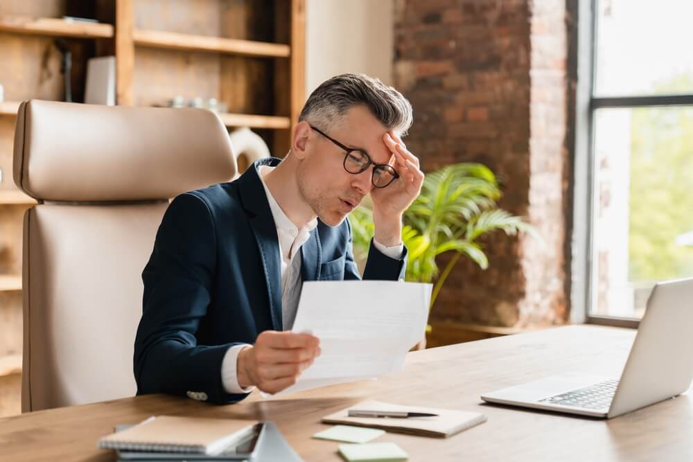 A business owner sitting in a chair at his desk with his hand on his head, worried after receiving a lawsuit and without business lawsuit protection.