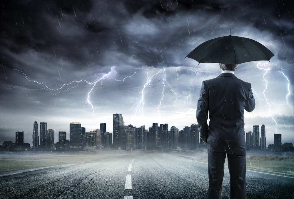 Businessman With Umbrella Looking Storm Over City, contemplating whether he needs umbrella insurance or liability insurance to protect his business.