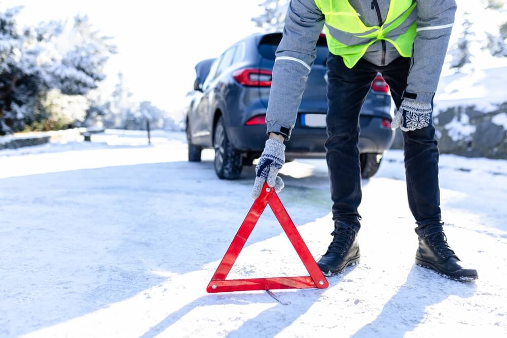 Driver placing a warning triangle on a snowy road after a car problem, emphasizing the value of holiday travel insurance tips for winter conditions.