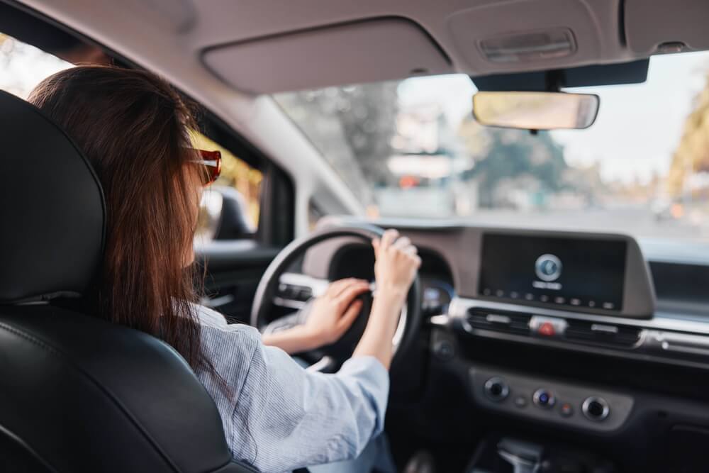 A woman drives through a sunny street while her car’s digital dashboard displays trip information, illustrating how telematics auto insurance connects everyday driving behavior with smarter and more adaptive insurance options.