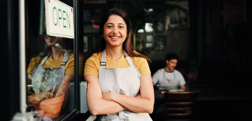 Small business owner smiles while standing at the entrance of her store with an open sign, highlighting independence and security backed by small business insurance.