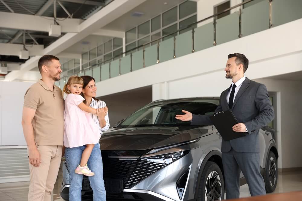 Salesman with clipboard showing new car to happy family in dealership.