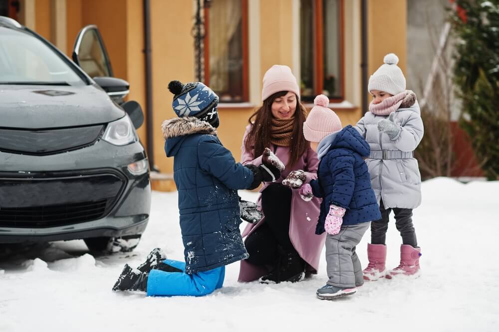 Young mother play with kids against electric car in the yard of her house at winter