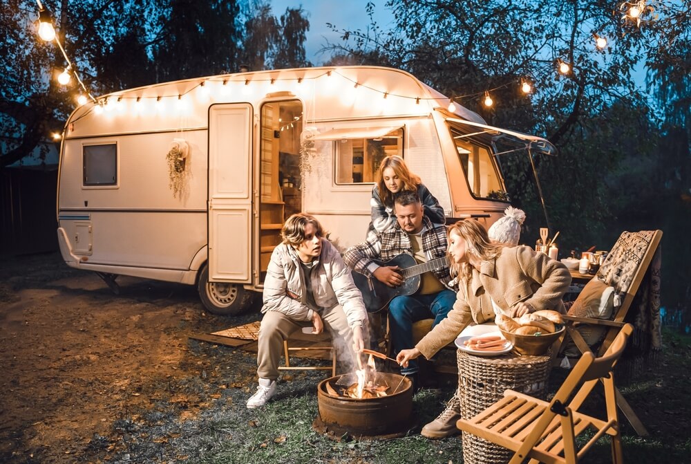 Family trailer travel. Children, brother sister, mom dad playing guitar, singing song at fire. Evening picnic in nature. Holiday barbecue BBQ food. Vacation weekend dinner. Road lunch. Camper, house on wheels.