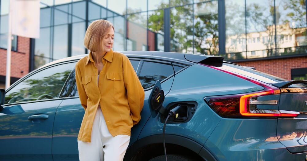 Woman standing beside a charging electric car outside an office building, highlighting electric vehicle insurance
