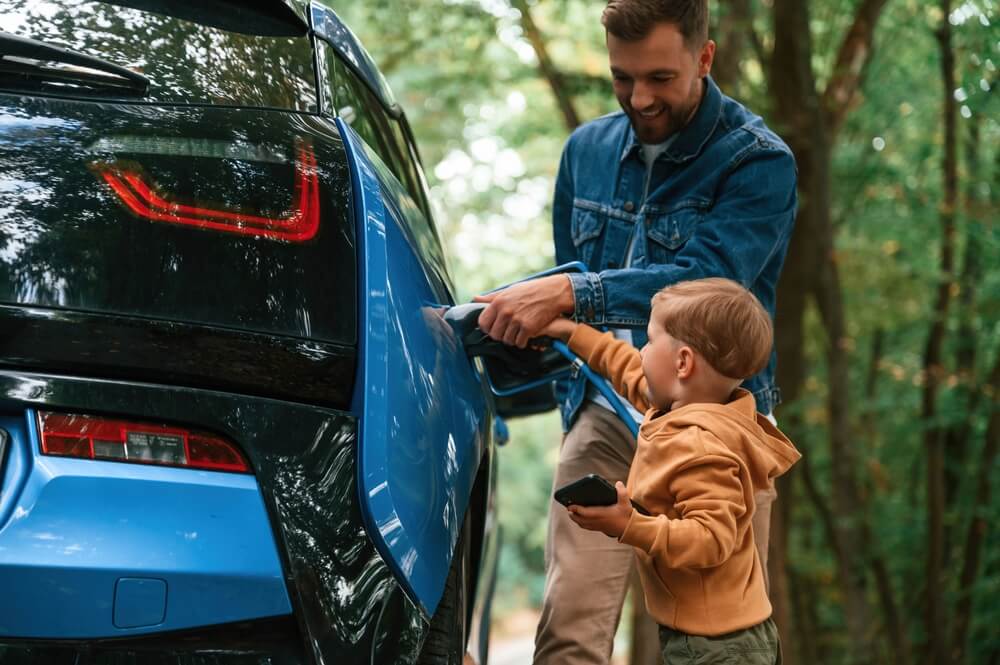 Man helps a child plug in an electric car charger in a wooded area, highlighting electric vehicle insurance.