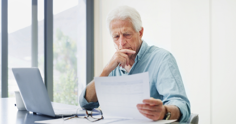 Older man reviewing financial documents at a table with a laptop, representing retirement planning and protecting home assets.