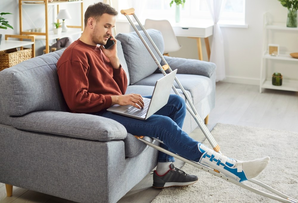 Employee with an injured leg using a laptop and phone at home, illustrating how disability insurance can support employees.