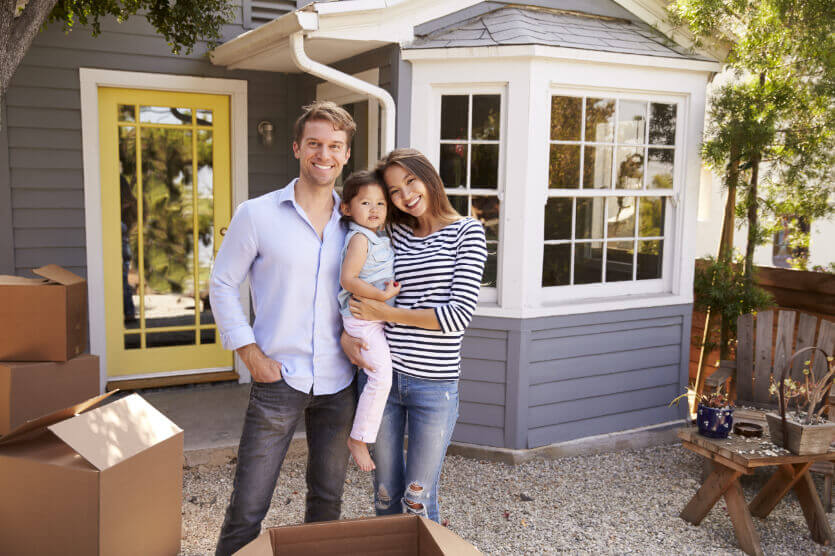 Happy mother, father, and small daughter standing in front of their rented home with moving boxes
