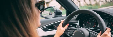 Close up of woman wearing sunglasses with hands on the steering wheel of a car.