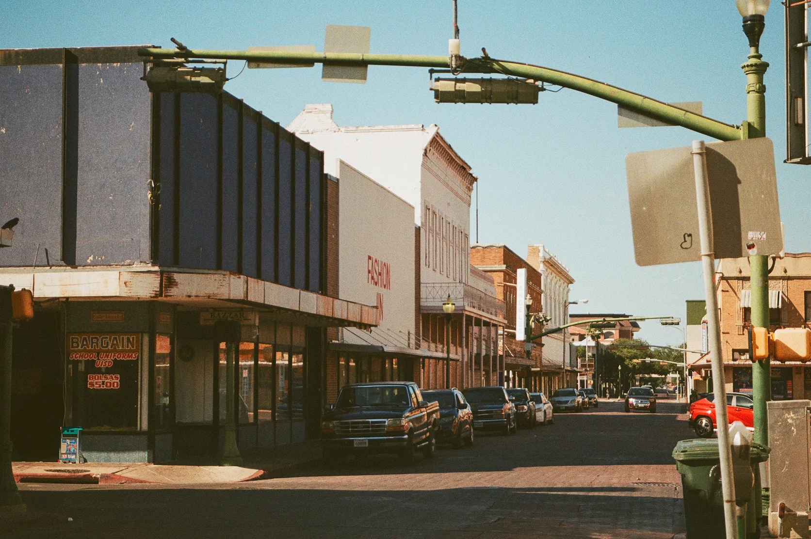 A film photograph of downtown Laredo with cars parked in front of storefront shops under the clear blue sky.