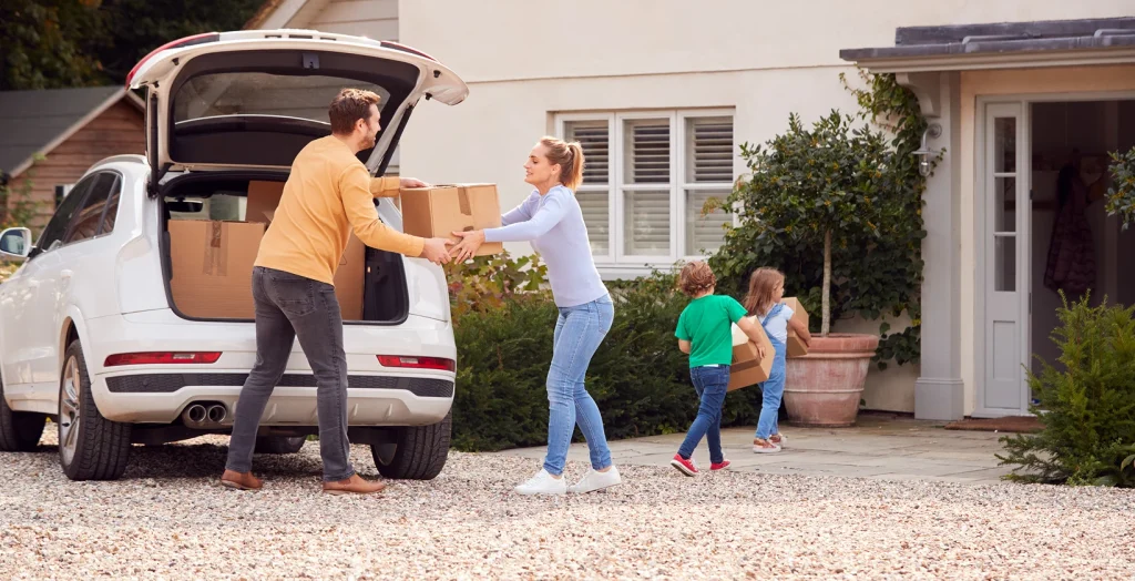 Mid-length shot of a family standing with luggage beside open boot of a parked car on the driveway in front of the house, concept of bundling auto and home insurance for savings.