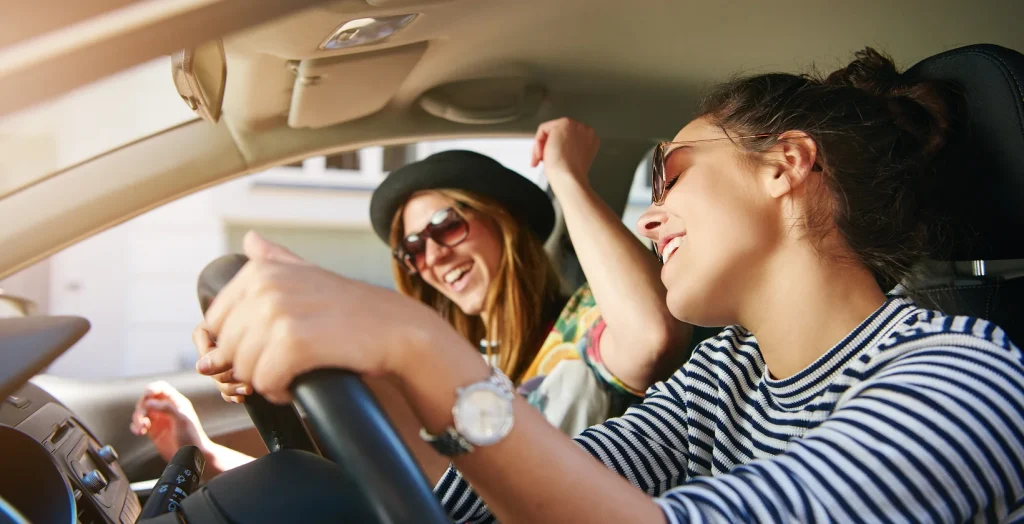 Two trendy attractive young woman singing along to the music as they drive along in the car through town viewed through the open side window, happy for the best car insurance with bundled discount.
