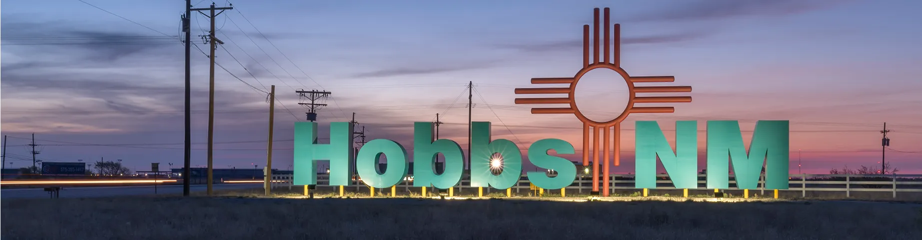 This photograph is an early morning shot of the entrance sign for Hobbs, New Mexico as seen from highway number 62. It is illuminated and contains the rising sun in the background.