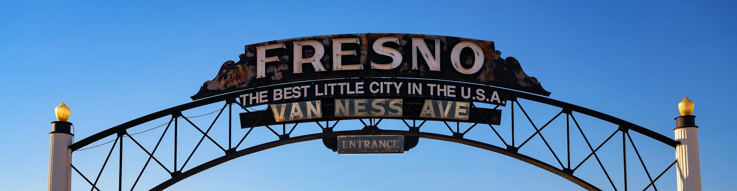 Rustic archway sign reading 'Fresno - The Best Little City in the U.S.A. over a quiet Van Ness Avenue at sunset.