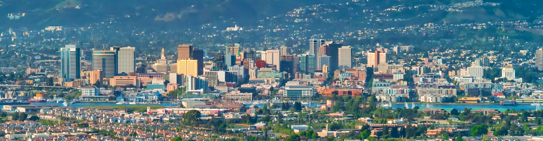 Aerial view of Oakland, California, showing the downtown skyline, surrounding residential neighborhoods, waterfront marina, and the East Bay hills in the background on a clear day.