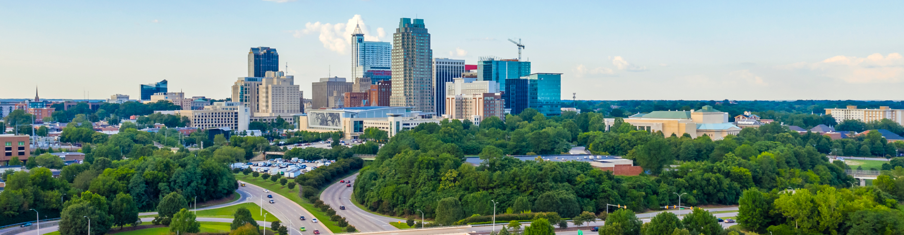 Aerial view of Raleigh, North Carolina, featuring the downtown skyline in the distance, surrounded by lush green trees, parks, and major highway interchanges under a bright blue sky with scattered clouds.