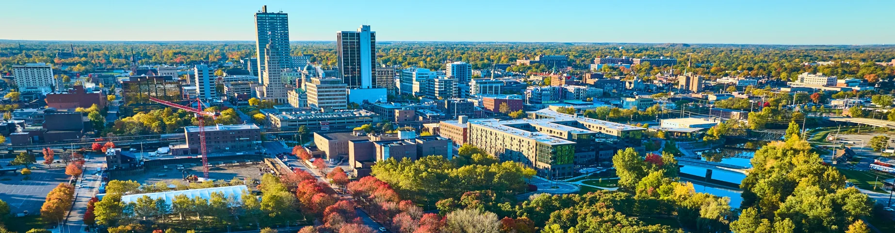Aerial view of downtown Indianapolis, Indiana, featuring a tree-lined park with colorful fall foliage, surrounding green spaces, and the city skyline under a clear blue sky.