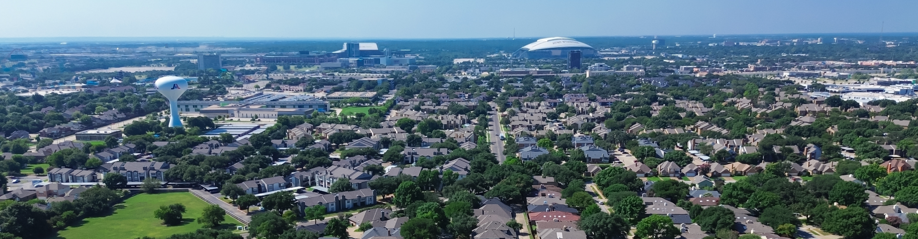 Aerial view of Arlington, Texas, featuring suburban neighborhoods, tree-lined streets, parks, and the city's skyline with AT&T Stadium visible in the distance under a clear blue sky.