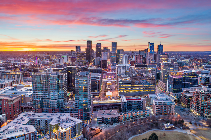 Aerial view of downtown Aurora, Colorado, at sunset, showcasing the city skyline, modern high-rise buildings, residential neighborhoods, and colorful evening sky.