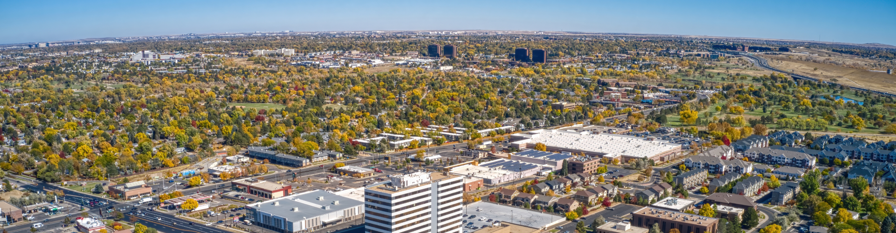 Aerial view of Aurora, Colorado, showing residential neighborhoods, office buildings, shopping centers, and tree-lined streets with colorful fall foliage under a clear blue sky.