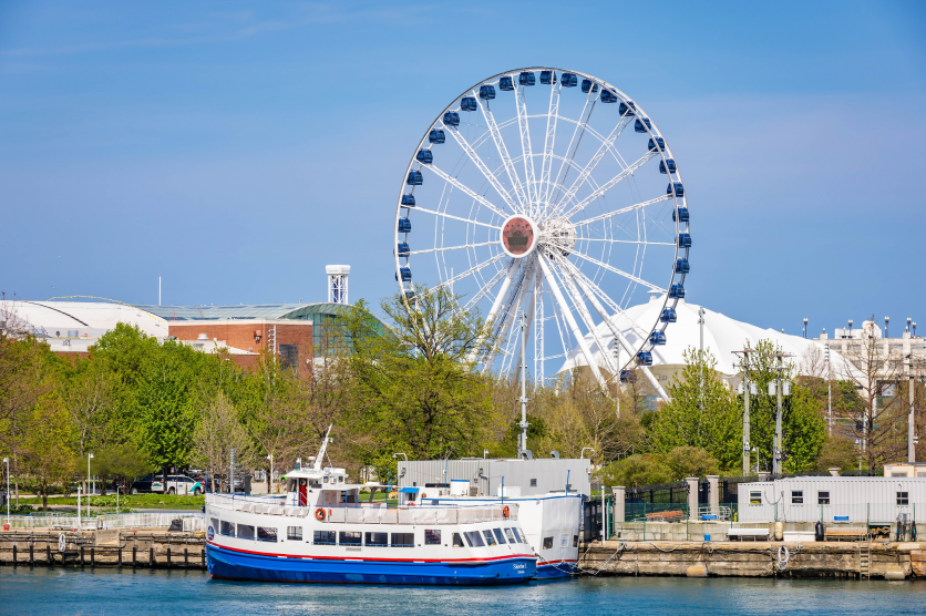 Waterfront view of Navy Pier in Chicago, Illinois, featuring the Centennial Wheel, a docked tour boat, and trees along Lake Michigan on a clear day.