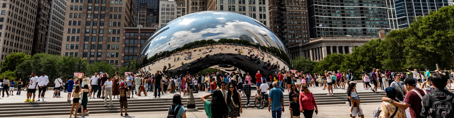 Cloud Gate ('The Bean') in Millennium Park, Chicago, Illinois, with visitors gathered around the iconic mirrored sculpture against the city's downtown skyline on a sunny day.