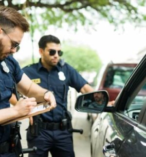 Police officers issuing a fix-it ticket to a driver during a roadside traffic stop, with one officer writing on a notepad beside the vehicle.