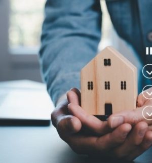 Hands holding a small wooden house with insurance checklist graphics, helping explain understanding replacement cost vs. actual cash value in home insurance.