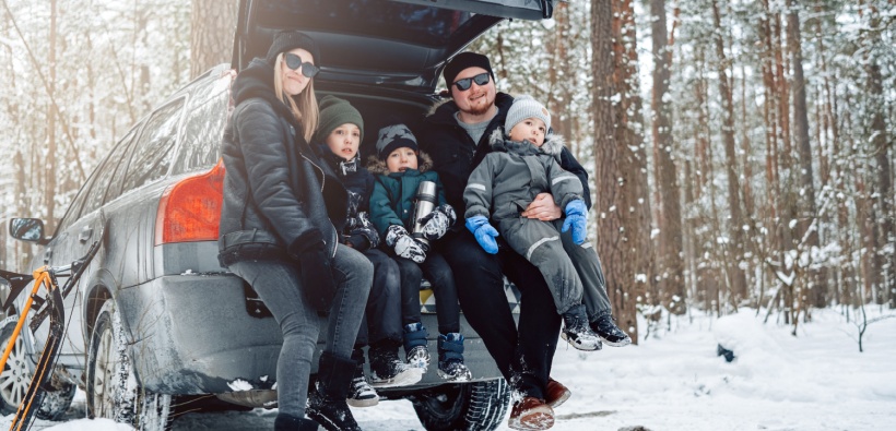 Young family sitting on their car trunk in a winter forest, highlighting the need for smart holiday travel insurance tips during cold-weather trips
