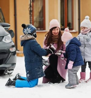 Young mother play with kids against electric car in the yard of her house at winter