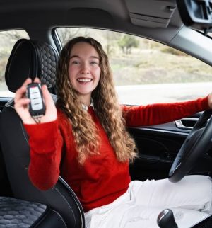 Young friendly positive woman holding a key of new white modern car. Portrait of happy cheerful female driver of new automobile showing a key, looking at camera and smiling.